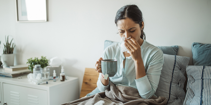 woman sick in bed, blowing nose with mug of tea in hand